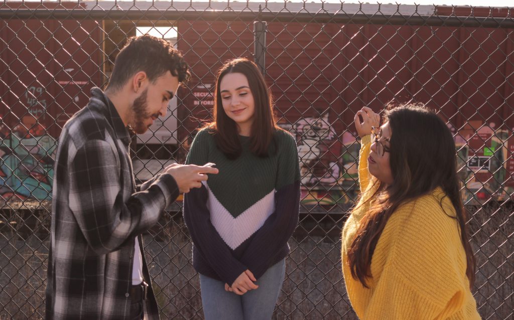 A young man in a check shirt and two young women in fashionable sweaters, all of different races, hang out by a fence in the sunshine, with a wall of graffiti behind them. The man is talking to the women while looking at his phone, the women appear to be interested in what he is looking at too. Earning the trust of a person, by establishing credibility with them, is essential to crossing our dividing barriers and becoming friends.