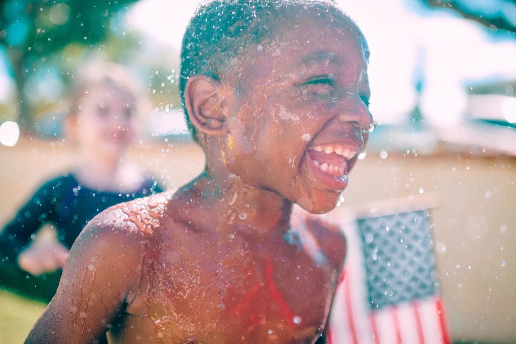 A young boy plays in water on a hot summer's day. He is smiling widely as a little girl stands behind him holding the stars and stripes. Independence Day can help remind us of the ultimate freedom we enjoy through our faith in Jesus Christ.