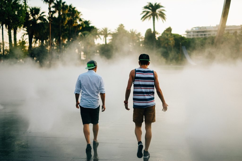 Talking about solutions is the final part of establishing trust and friendship with someone who is different to us. Finding a solution to a common problem is the greatest blessing we can enjoy from having a trusted relationship with someone different to us. In this photo, two young men in shorts and shirts, with backwards baseball caps, walk on slick concrete with what appears to be smoke obscuring the view of palm trees ahead of them. They are talking, with one of the men gesturing as he speaks.