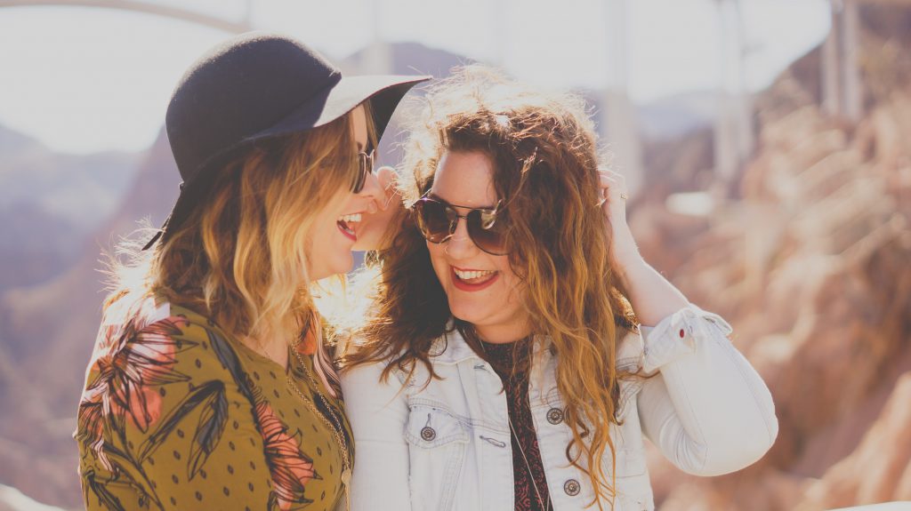 Two fashionable young ladies stand close together outside in the sunshine. They both wear sunglasses and are laughing. To help a friend along on their faith journey, you can invite them to attend your small group, or share your story with them.