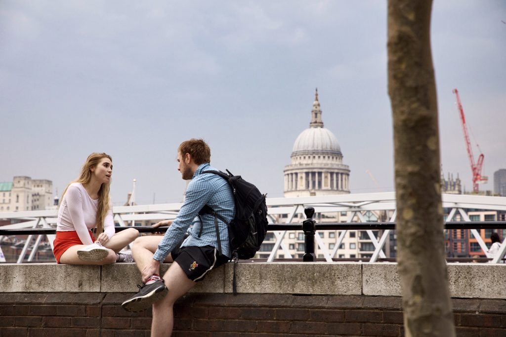 A young couple sit opposite each other on a wall by the River Thames in London. Teh sky behind them looks cloudy, but they both wear shorts and long-sleeve tops. We all have an opportunity to share Jesus with others, all it takes to start is a friendly conversation!