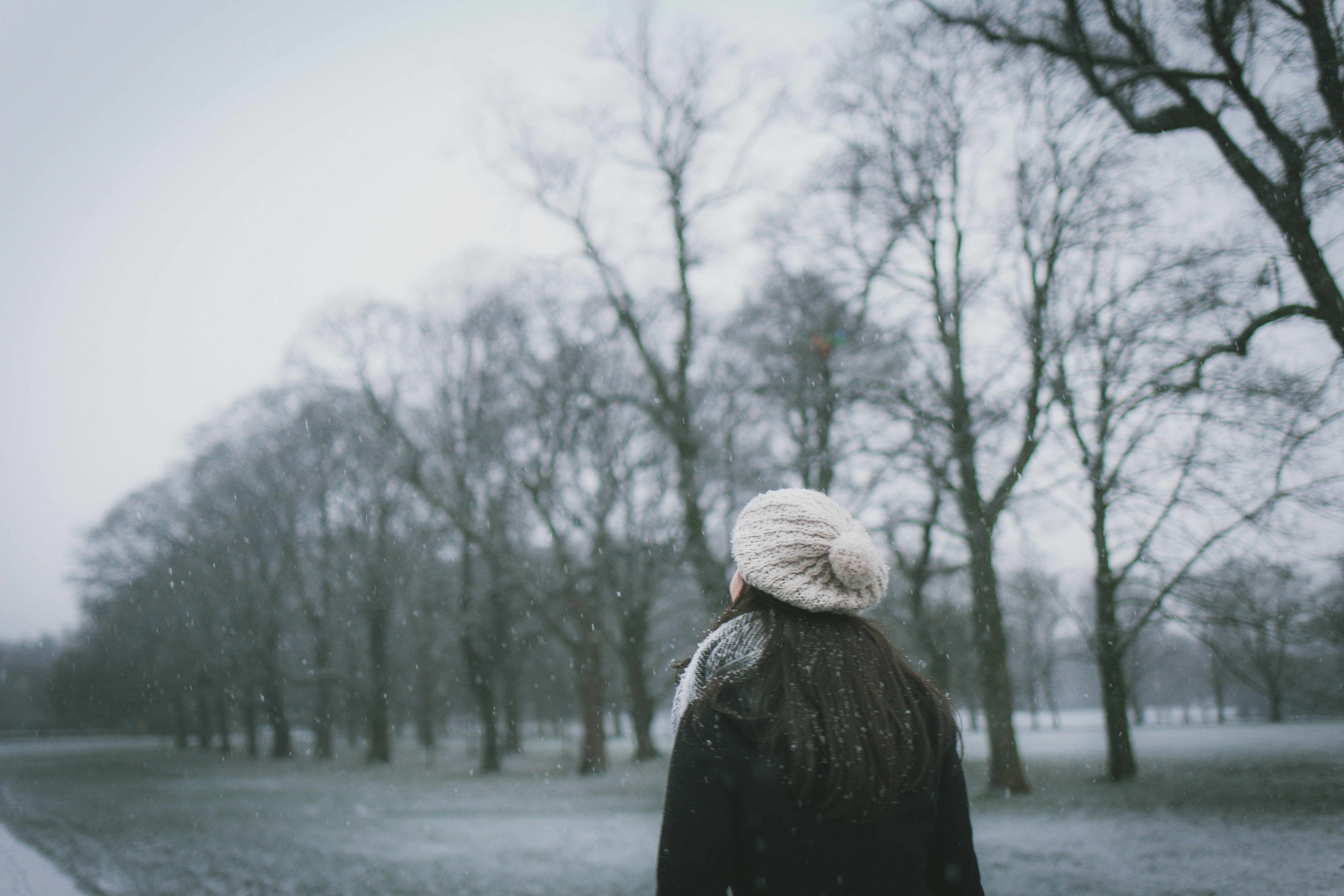 Young woman wearing black winter coat and white woolen hat looking up thoughtfully at gray sky with bare trees in background and snow falling.