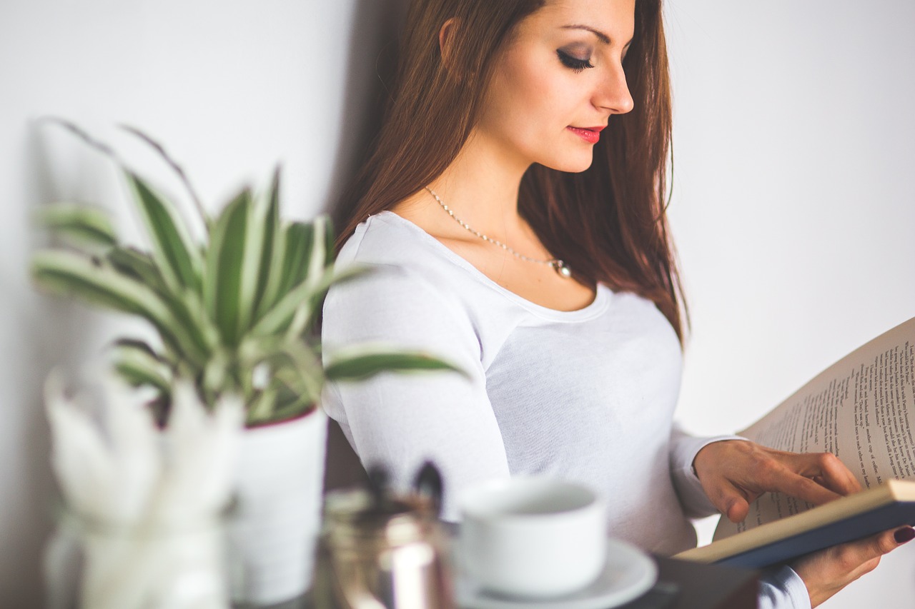 Woman with dark brown hair wearing white long sleeve shirt holding a book, relaxing against a wall with green plant and teacup in foreground.