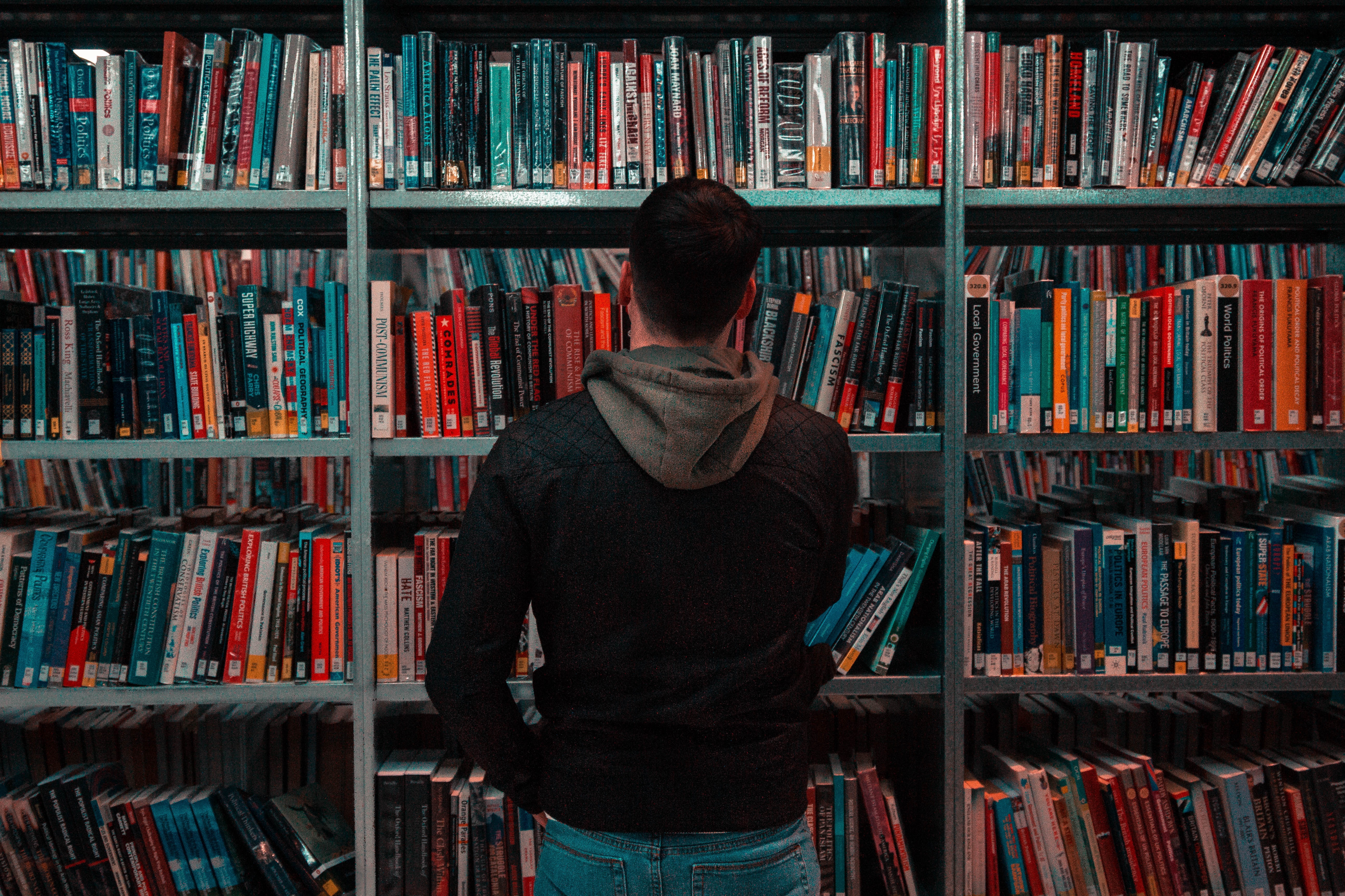 Young man wearing dark sweatshirt looking thoughtfully at library shelves of colorful books.