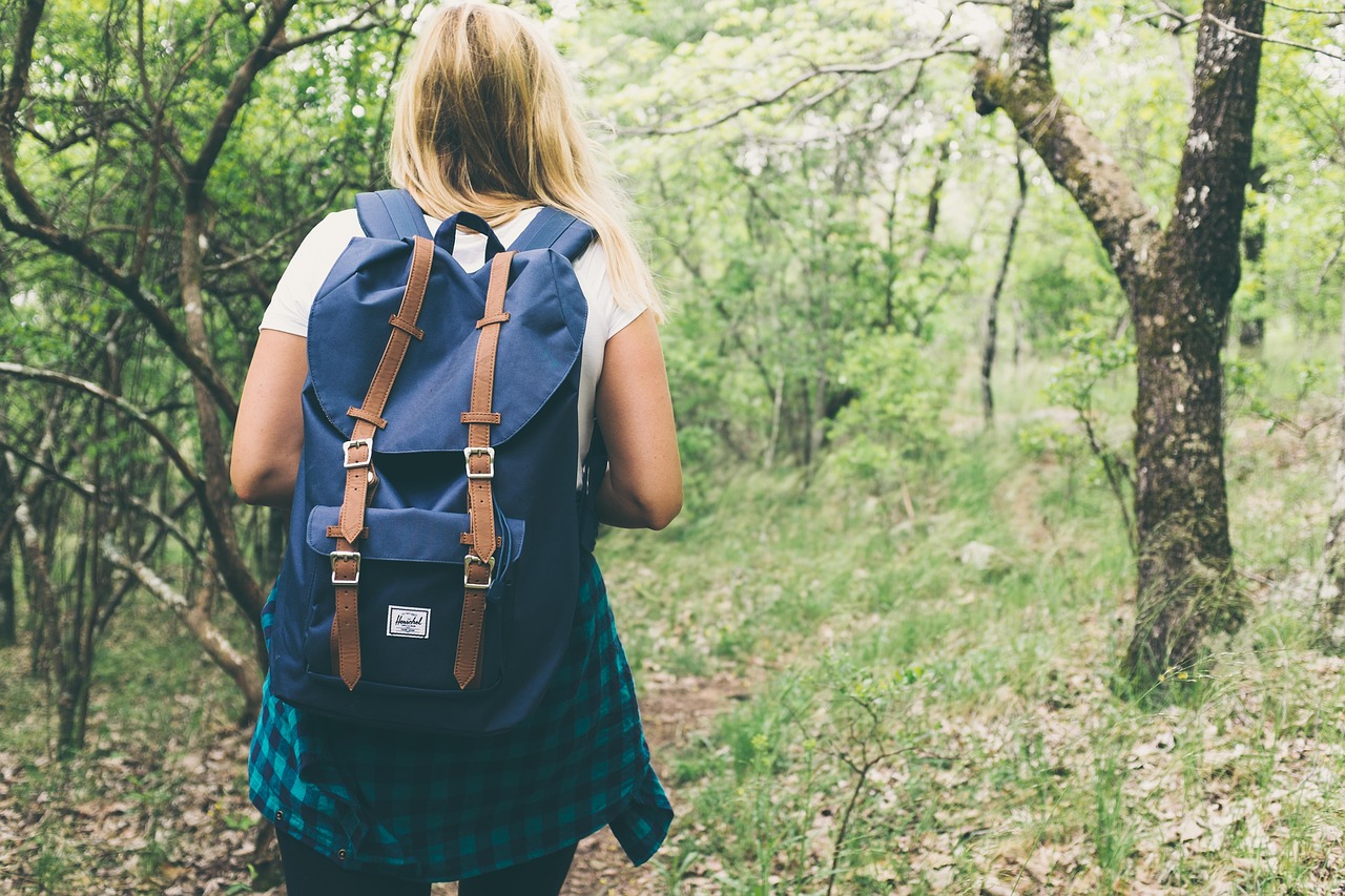 Young blonde woman walking down a wooded trail wearing short sleeves and a backpack