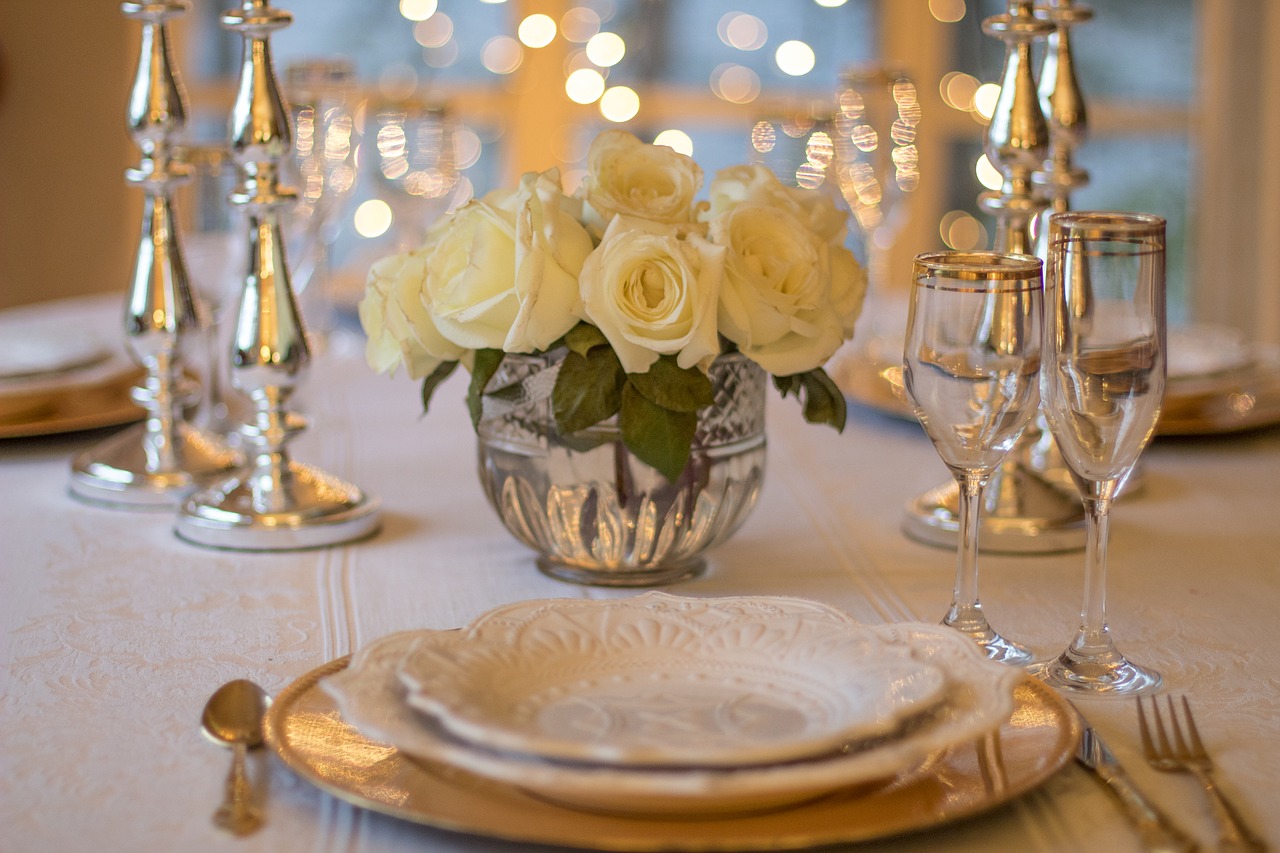 Elegantly set table with white tablecloth, fine china, silverware, silver candlesticks and lights in background, and a crystal vase of white roses