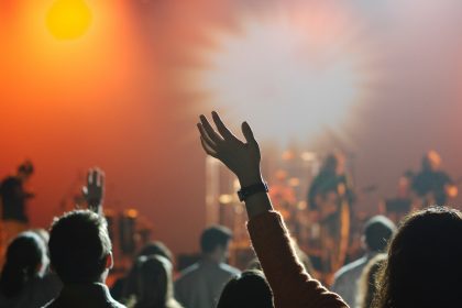 People raising hands at worship concert with warm, bright lighting in background and band on stage