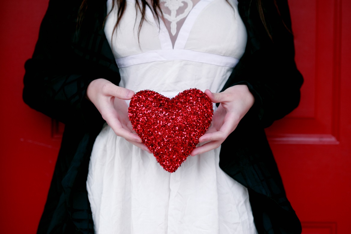 Young woman wearing a white dress with black sweater standing against red door holding sparkly red Valentine's heart