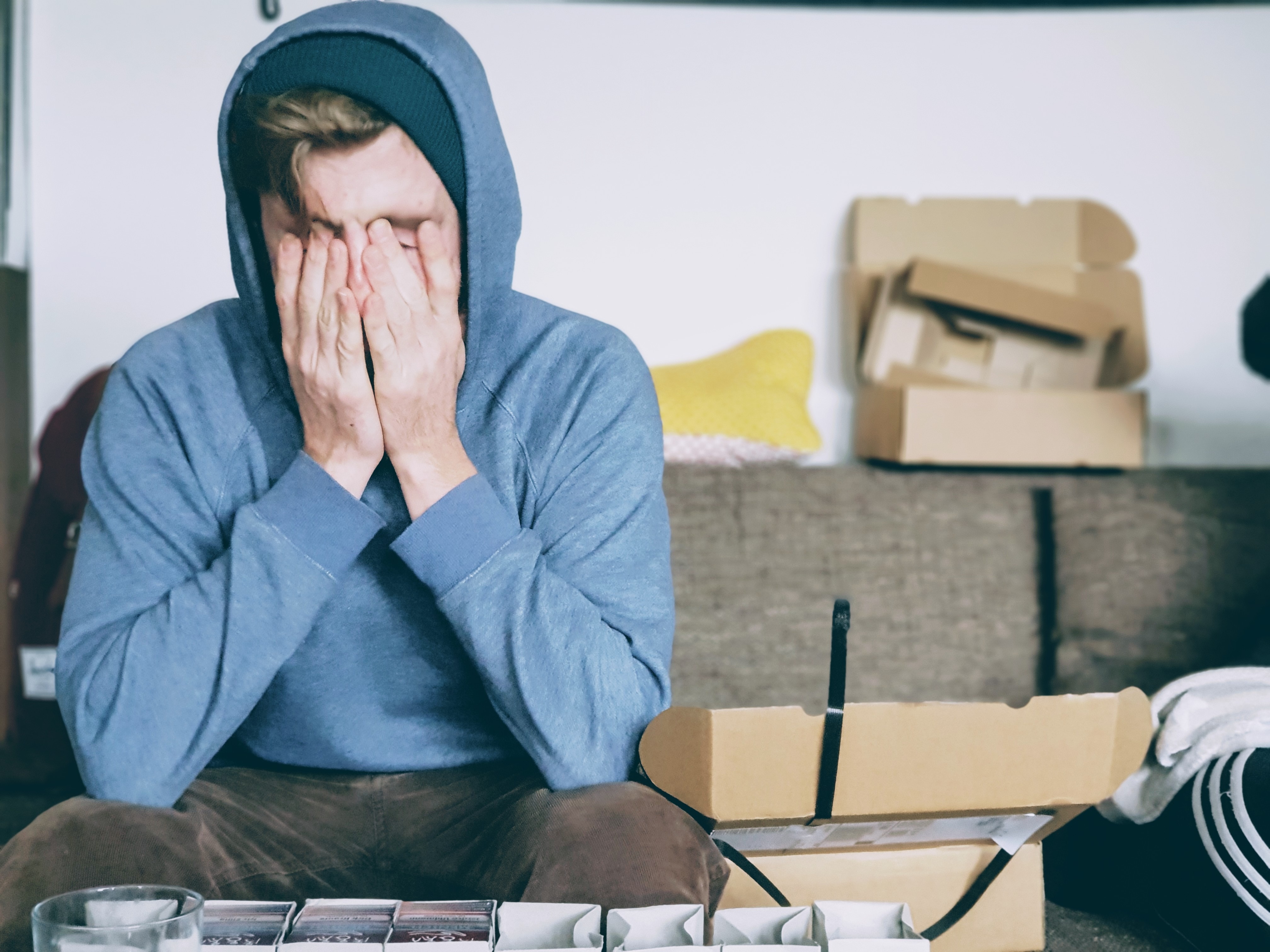 Young caucasian man wearing blue hoodie sitting on couch with face in hands, surrounded by empty cardboard boxes