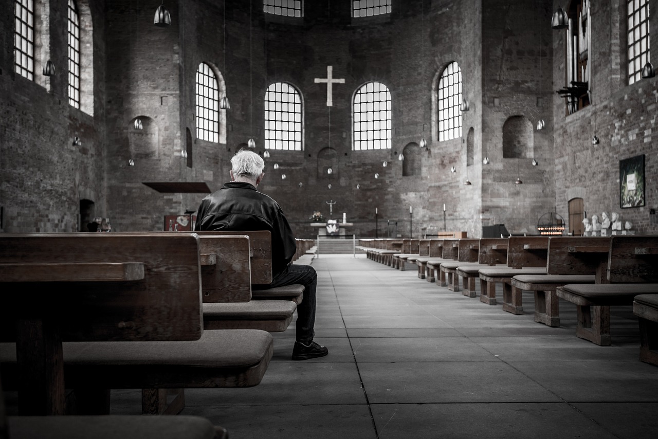 Elderly white-haired man sitting in pew inside a dark gray church