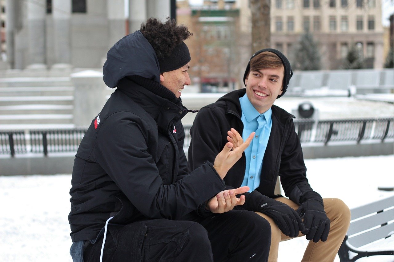 two young men wearing winter coats and hats sitting on a bench outside talking and smiling