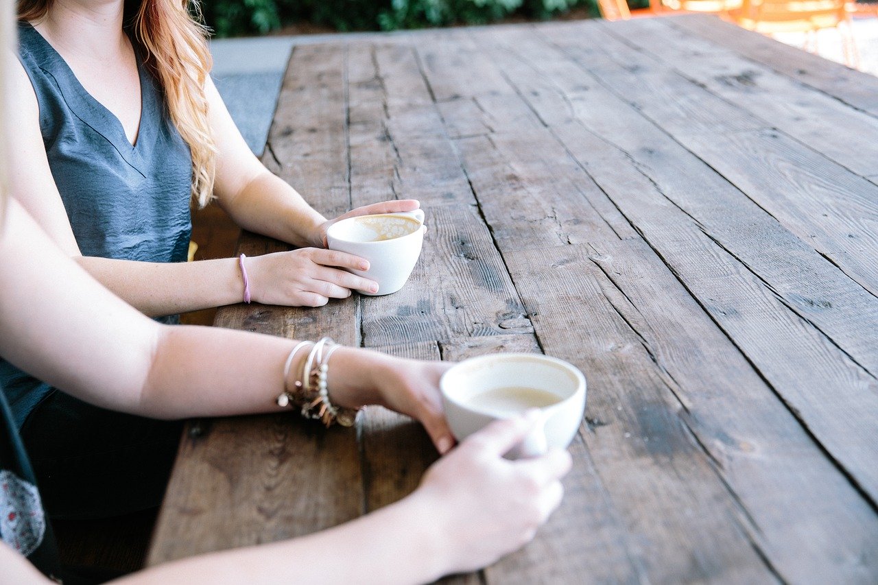 Two young women sitting at a wooden picnic table outside holding white coffee cups