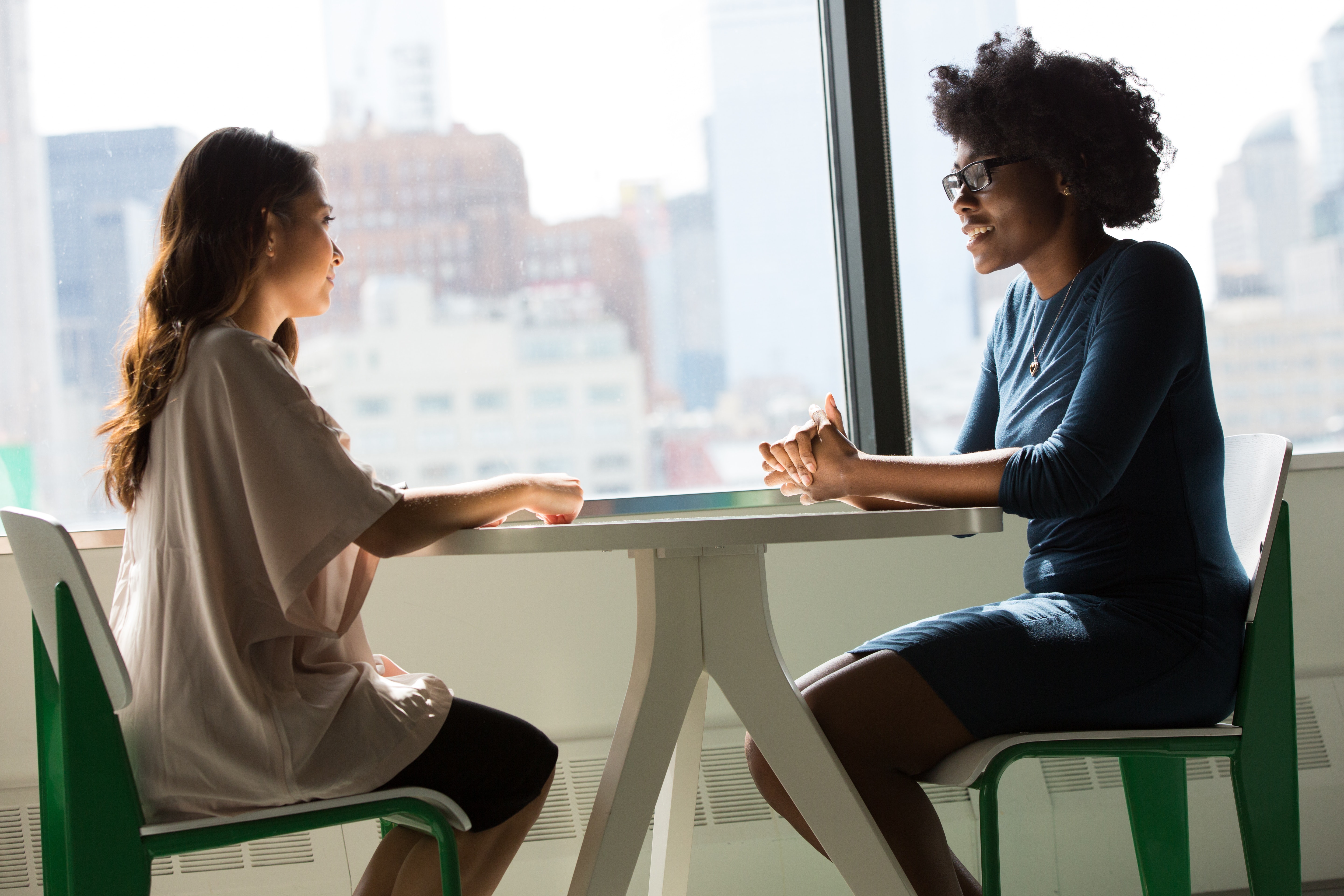 an african american woman sitting at a white table by a window talking with a young caucasian woman with dark hair