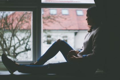 sad young woman quarantined sitting by window in a dark room looking outside