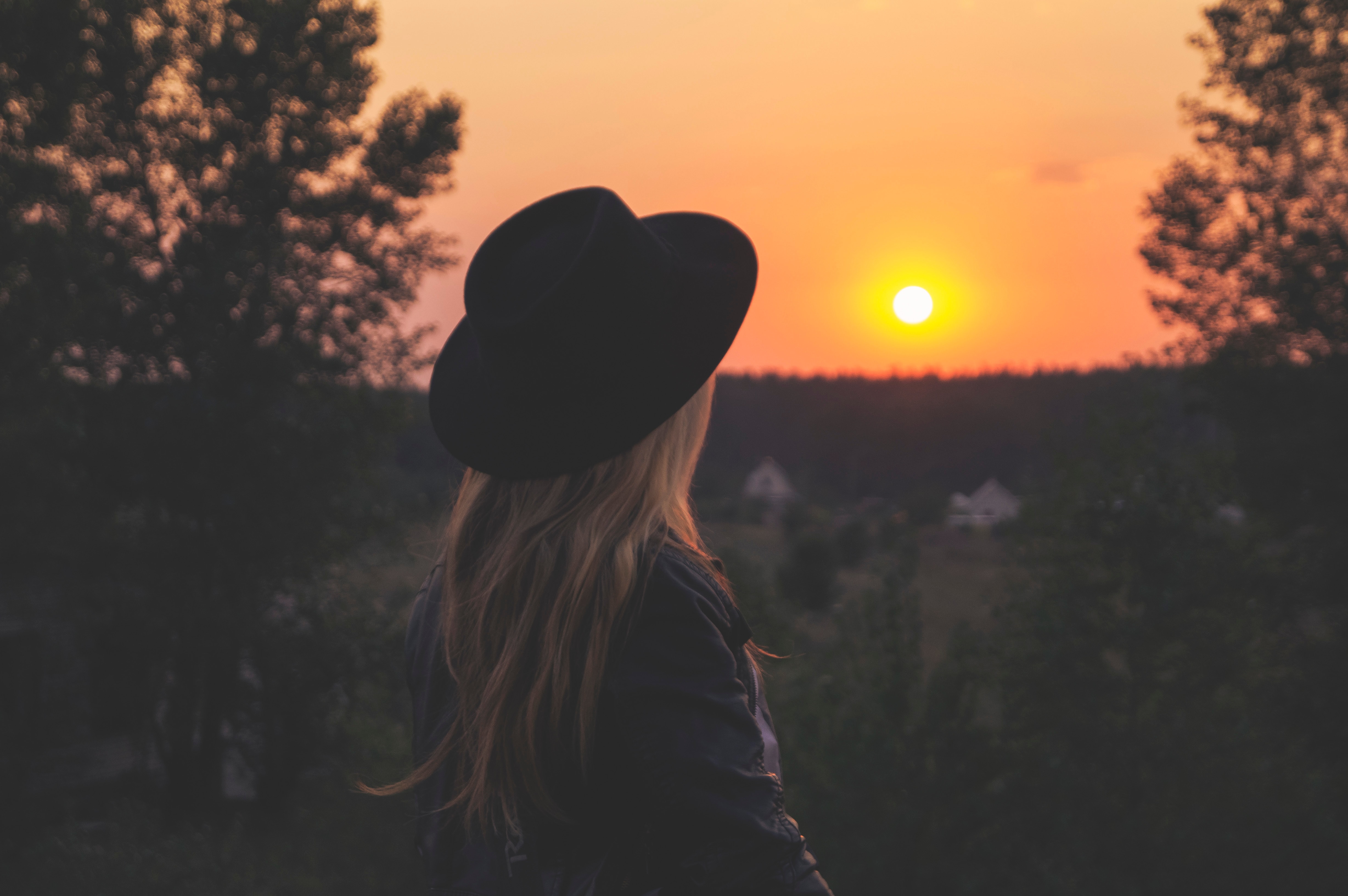 young blonde woman wearing black hat facing setting sun on horizon