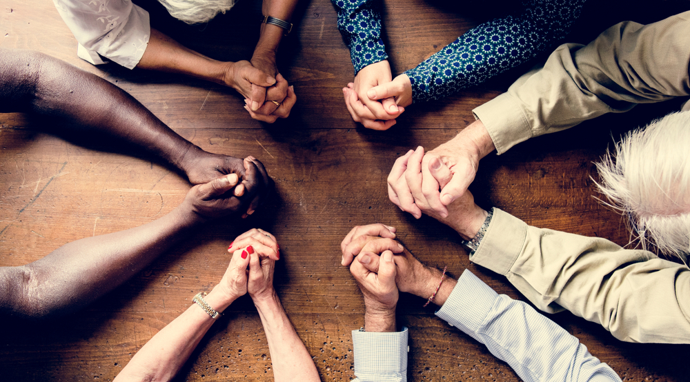 Birds-eye view of small group of diverse people with hands folded on a tabletop in prayer