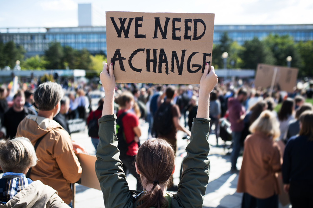 crowd of people protesting outside with young white woman holding a cardboard sign saying "we need a change"