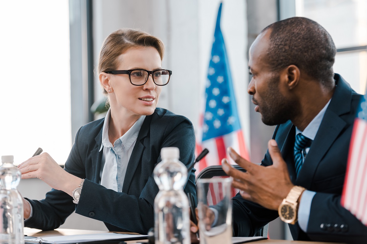 Black male and white female government officials talking cordially at a meeting with American flag in background