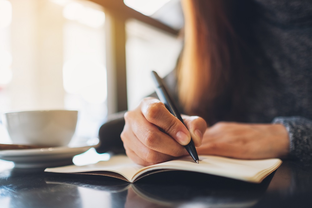 Close-up of young woman writing in a notebook with a cup of coffee on the table