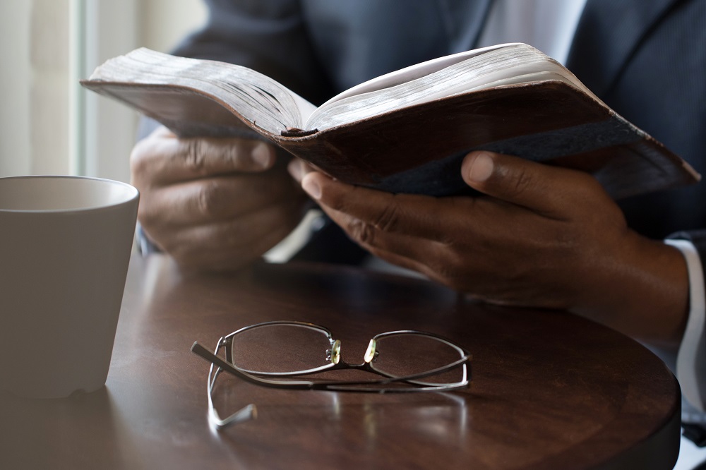 Close-up of black man reading book or Bible with white mug and eyeglasses on table