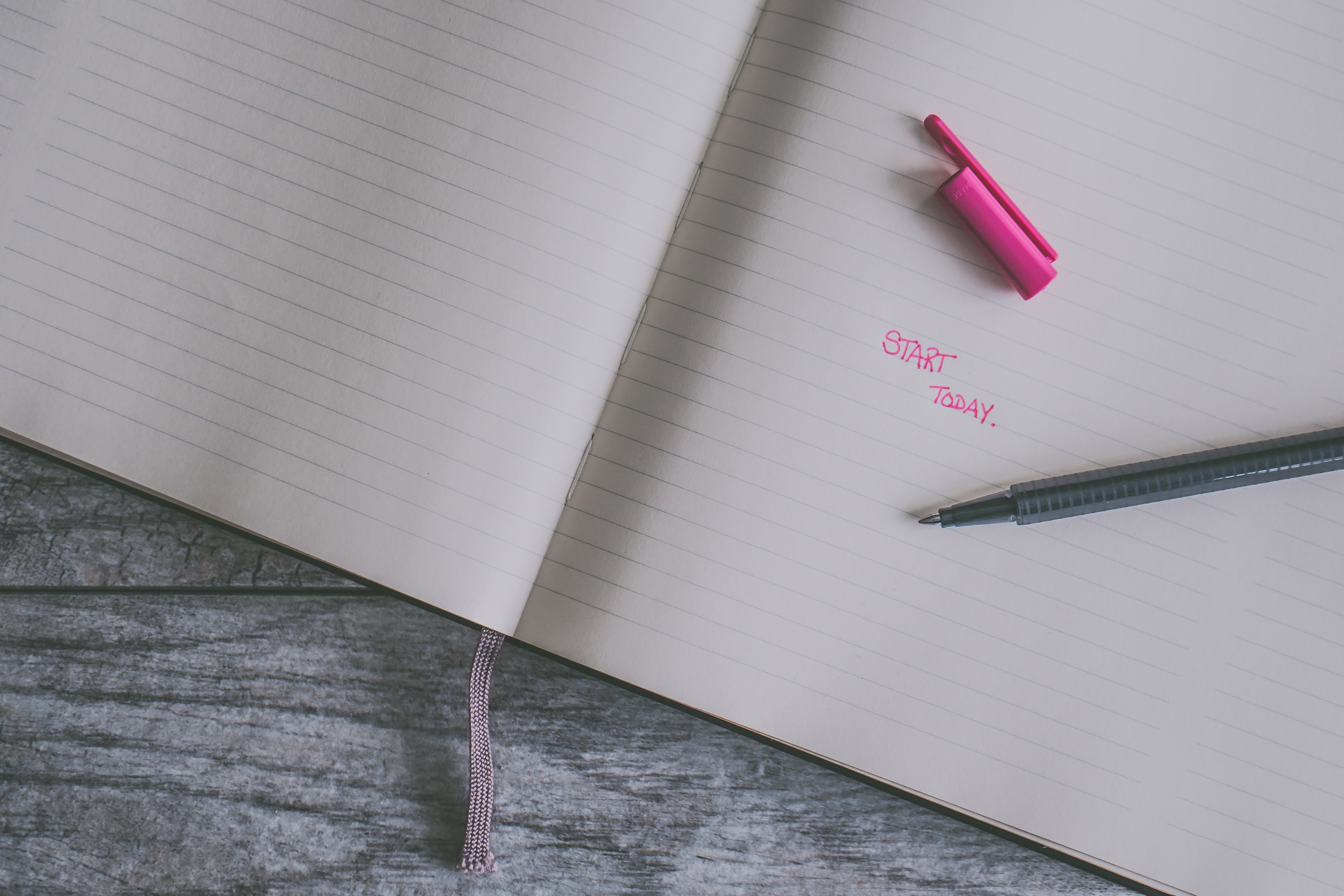 A plain notebook displays in pink ink the words written 'Start here'. Start now with your fresh beginning this year.