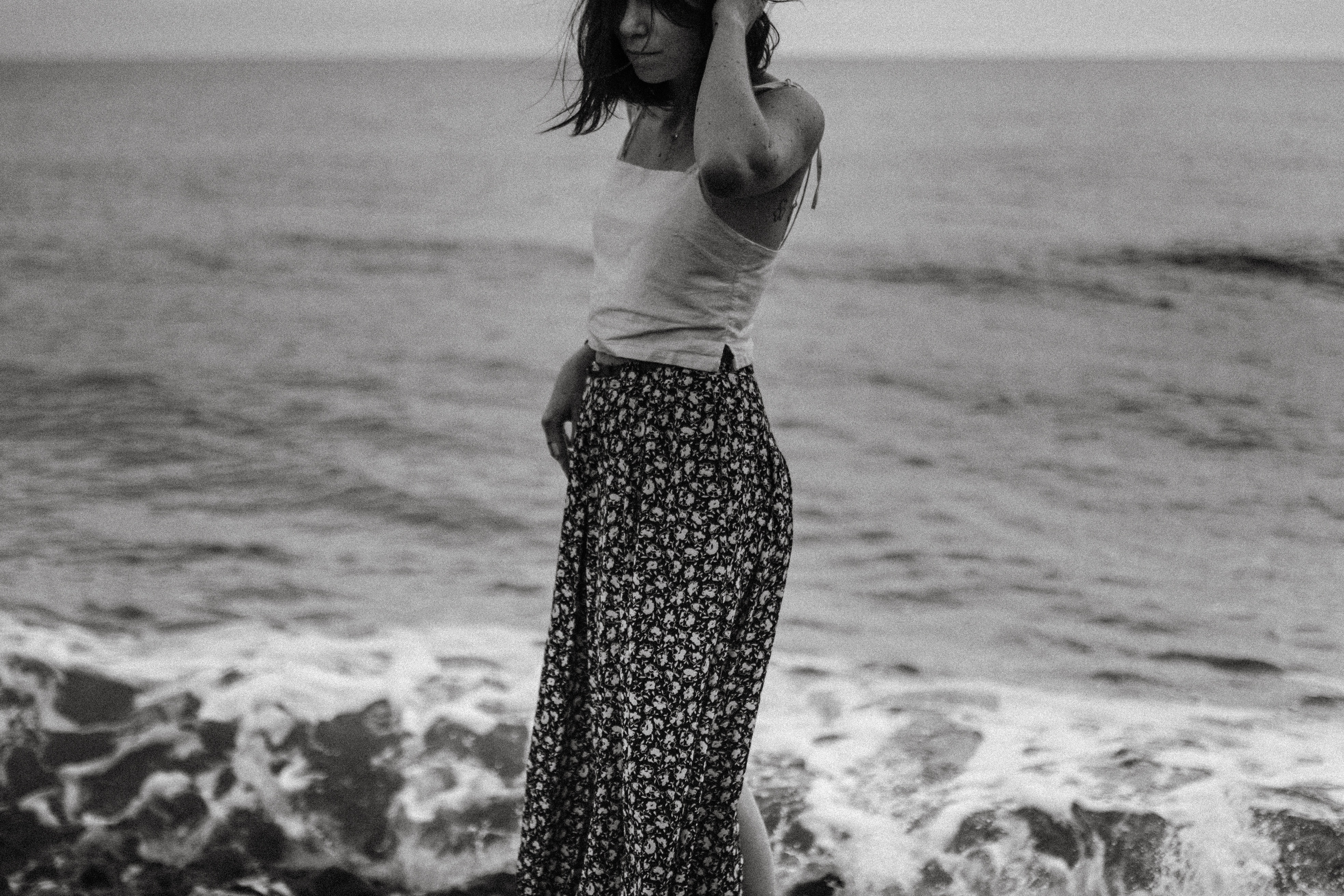 A young woman walks on a windy beach in a black and white photo, looking mournful.