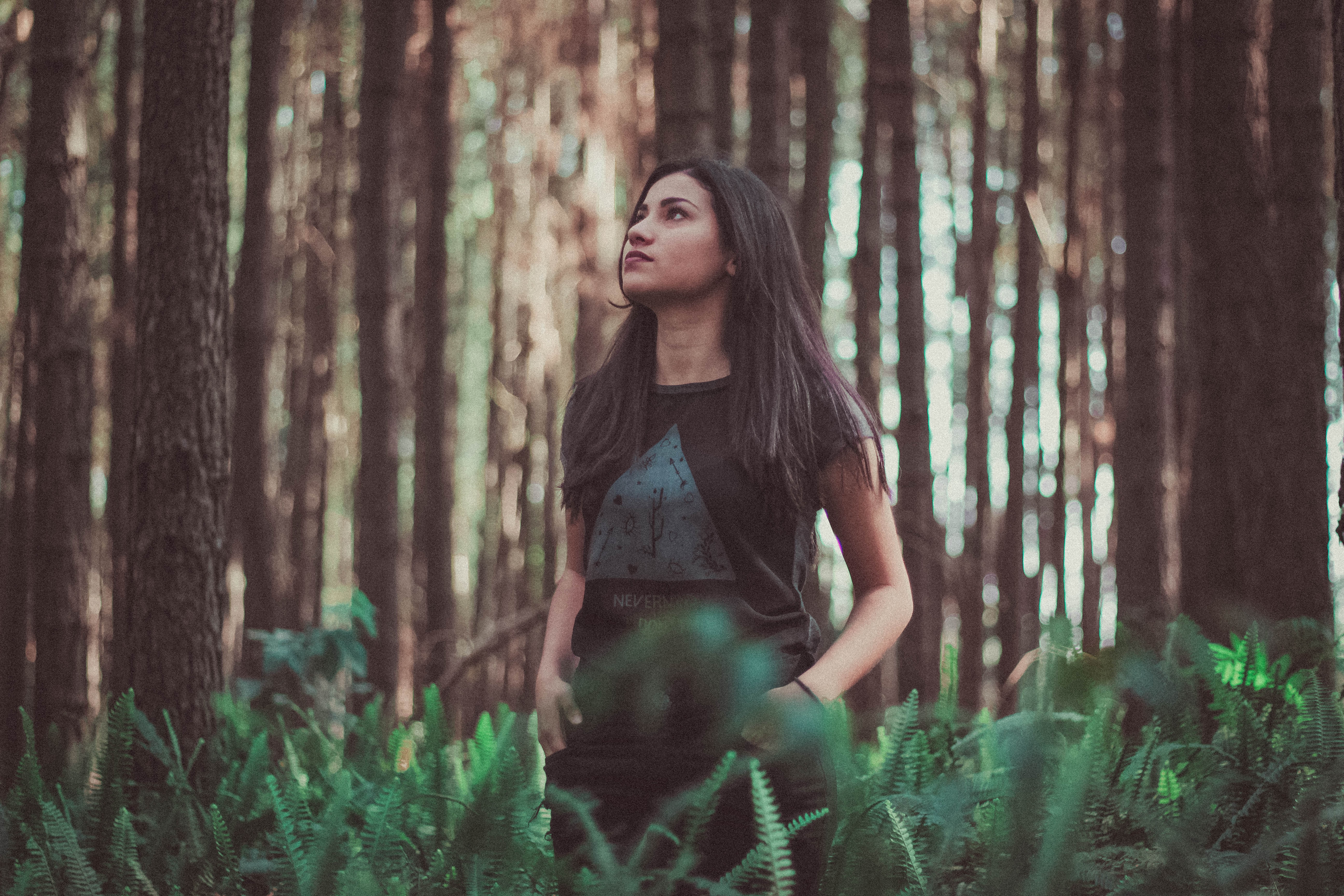 A young woman stands with her hands in her pockets, among ferns and trees in a forest. She is looking up towards the sky. She could be wondering if she can trust God. To her we would say yes!