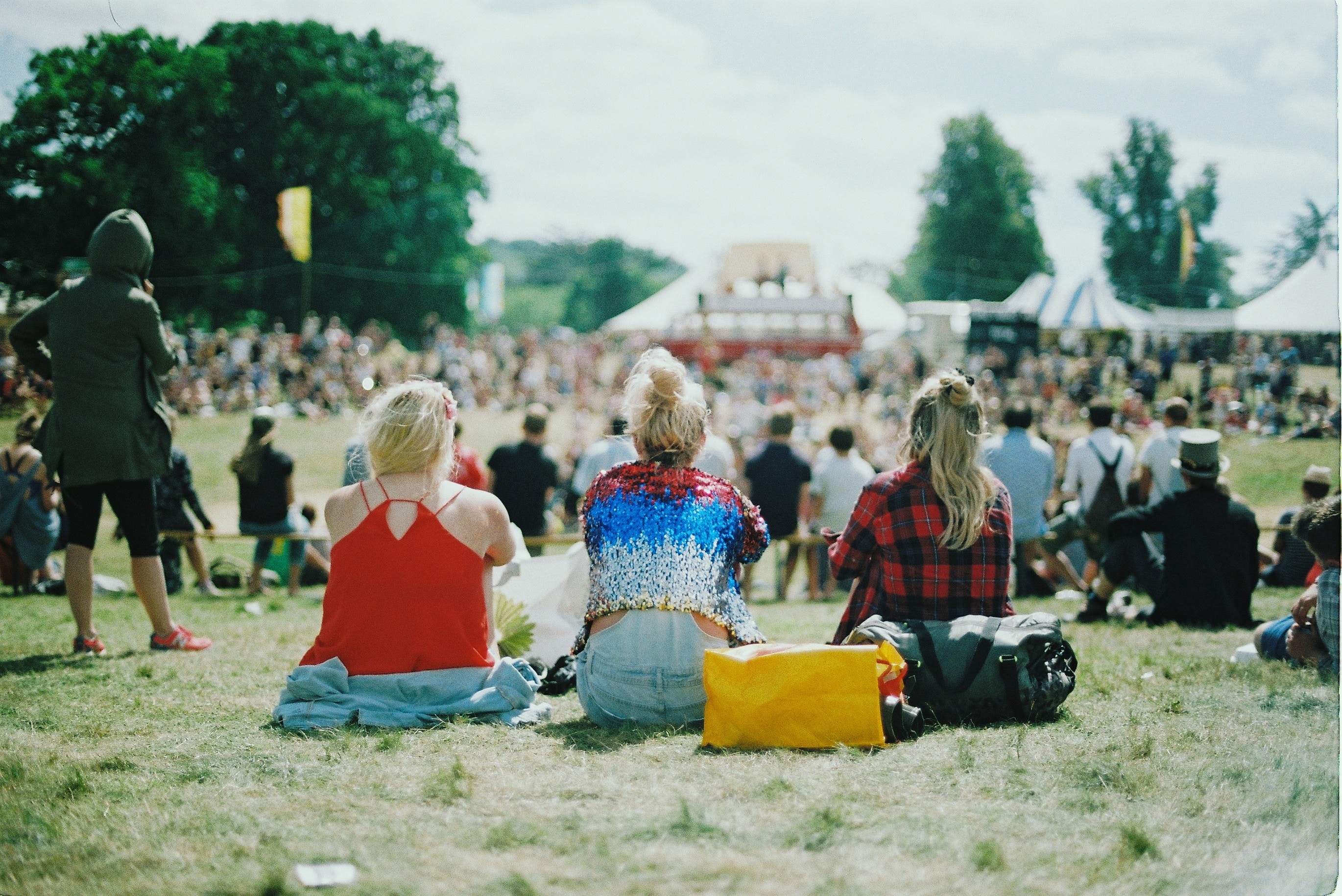 Three female friends sit on a field at a busy festival, with their backs to the camera. You can invite your non-believing friends to read the Bible with you and they, and you, will be blessed.