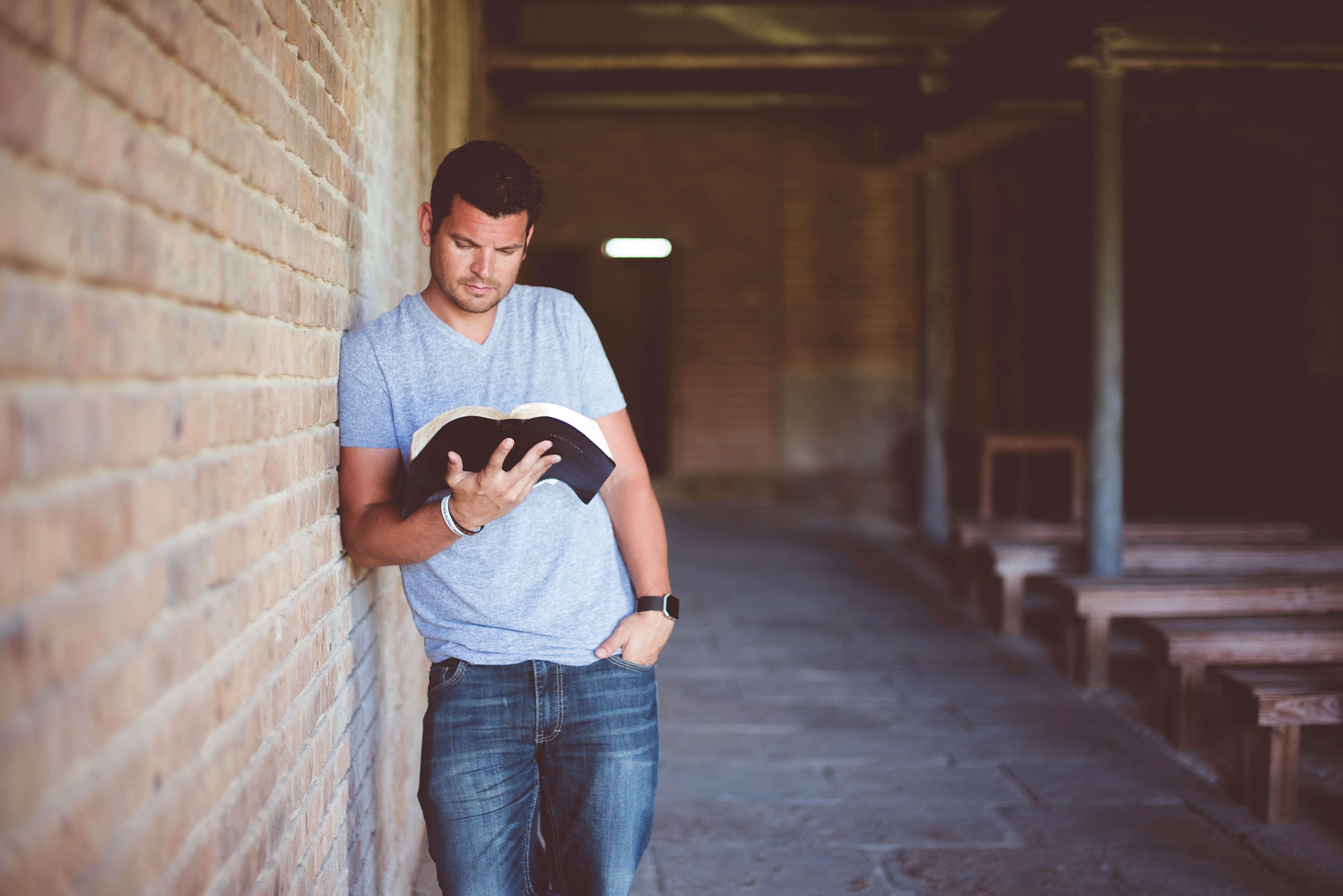 A man in jeans and t-shirt stands leaning against a wall as he reads his bible. Plain wooden benches are arranged in rows beside him. By reading our Bibles we can see all the ways in which we can in fact trust God.