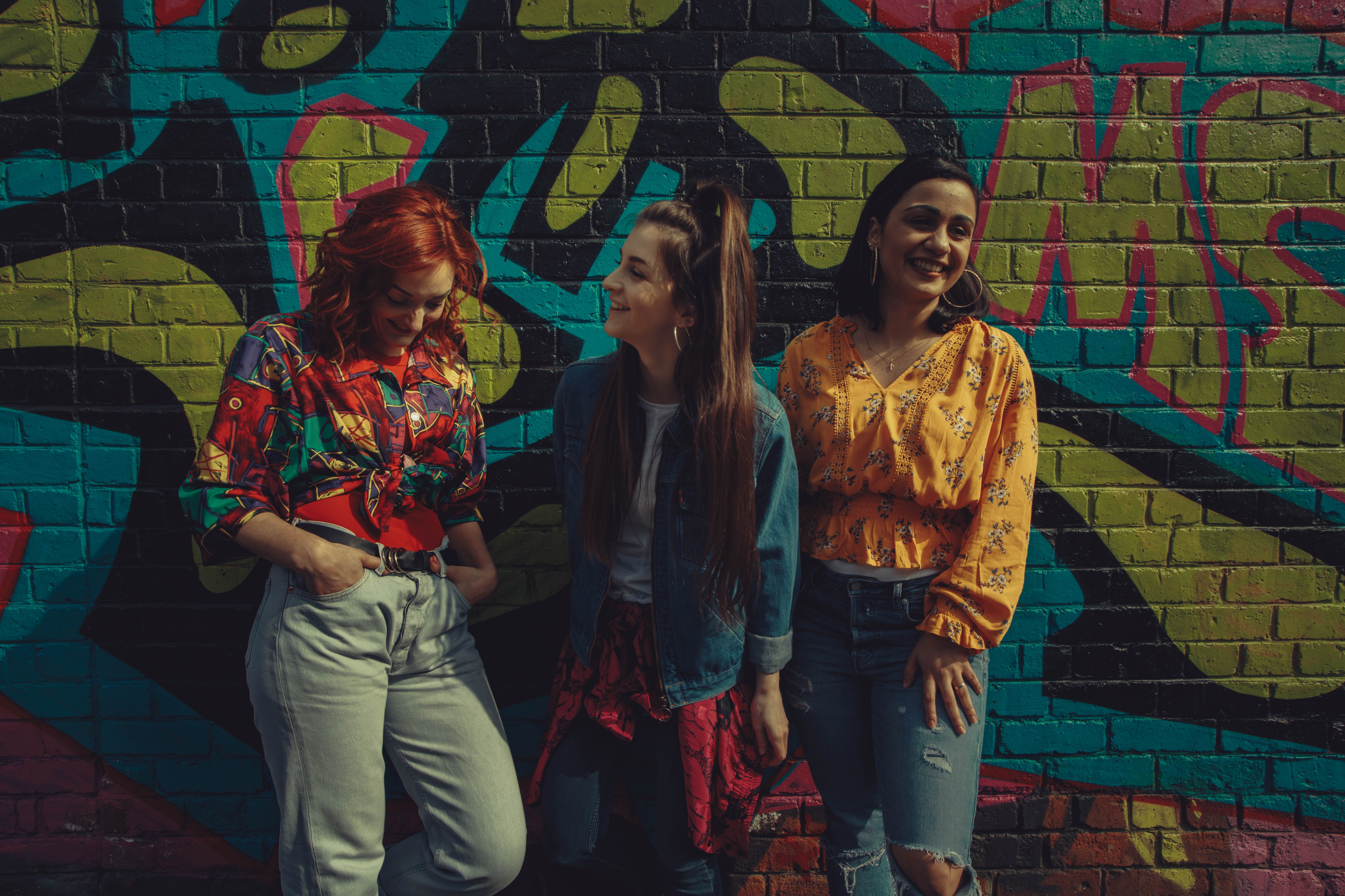 Three young women in colorful shirts and denim stand against a graffiti wall, they're all smiling. When you ask a non-believer friend to read the Bible with you, you can be sure that God's Word will speak to them in the way they personally need it to.