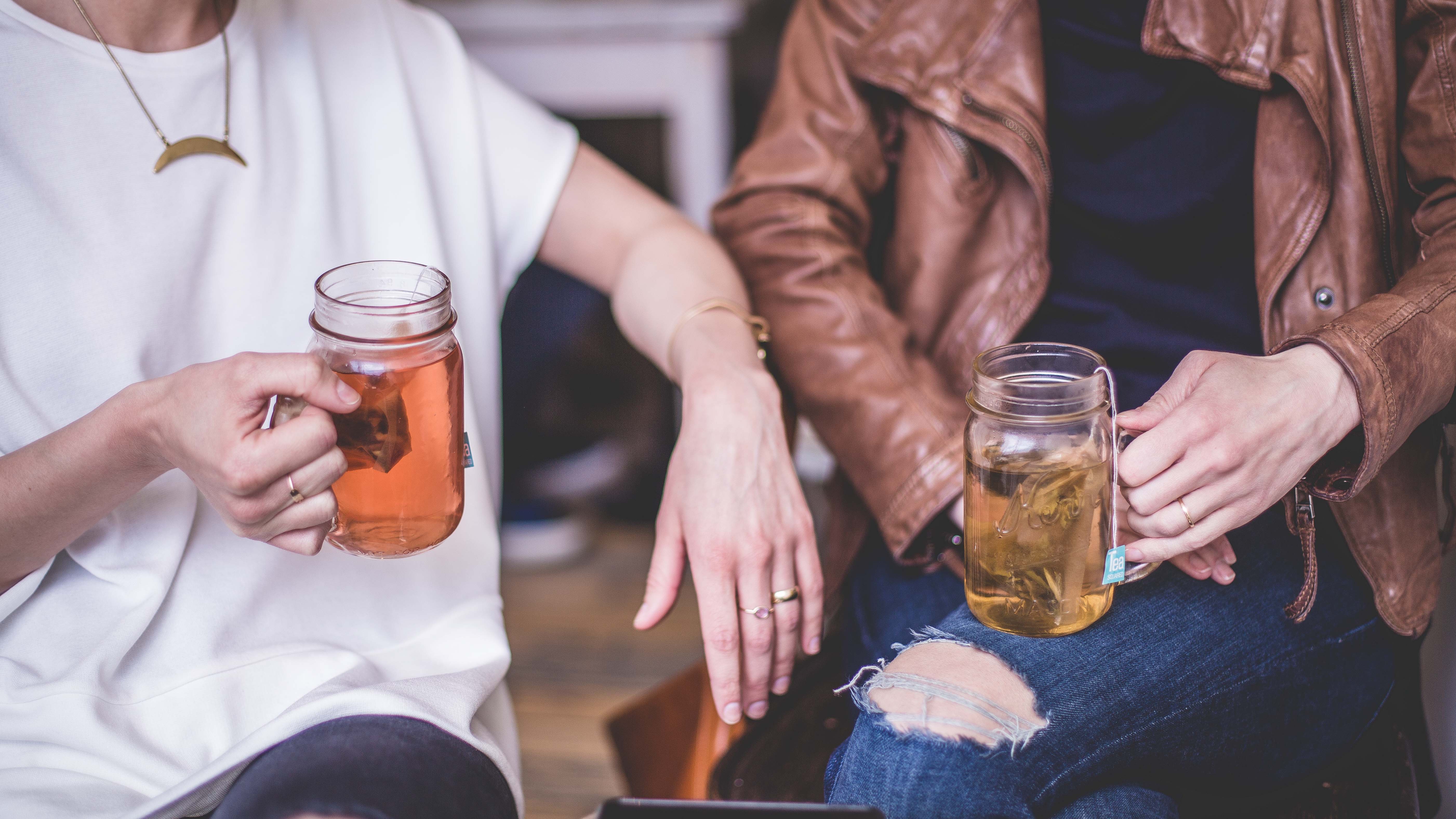 Two women holding mason jar mugs with tea, sit closely and chat. Creating a warm and friendly, non-judgemental environment can help your non-believer friends feel safe to open up and allow God to work in them as you read the Bible with them.