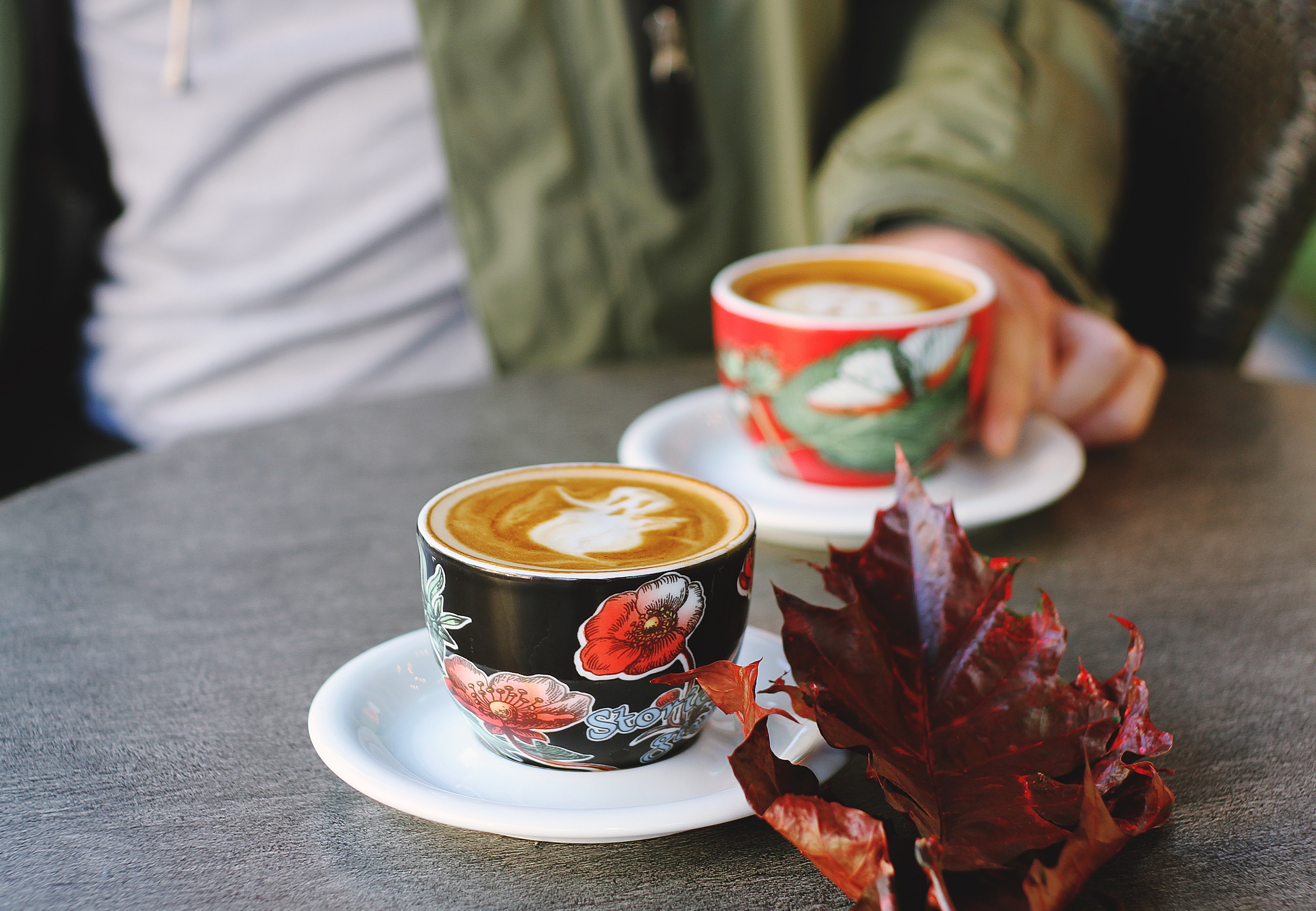 Two colorful cups on white saucers with on a table with a large red leaf and are filled with fresh coffee. A person sits behind the far mug, holding it delicately in the saucer. There are practical considerations for your meeting with a non-believer friend to read the Bible, this article gives you some tips!