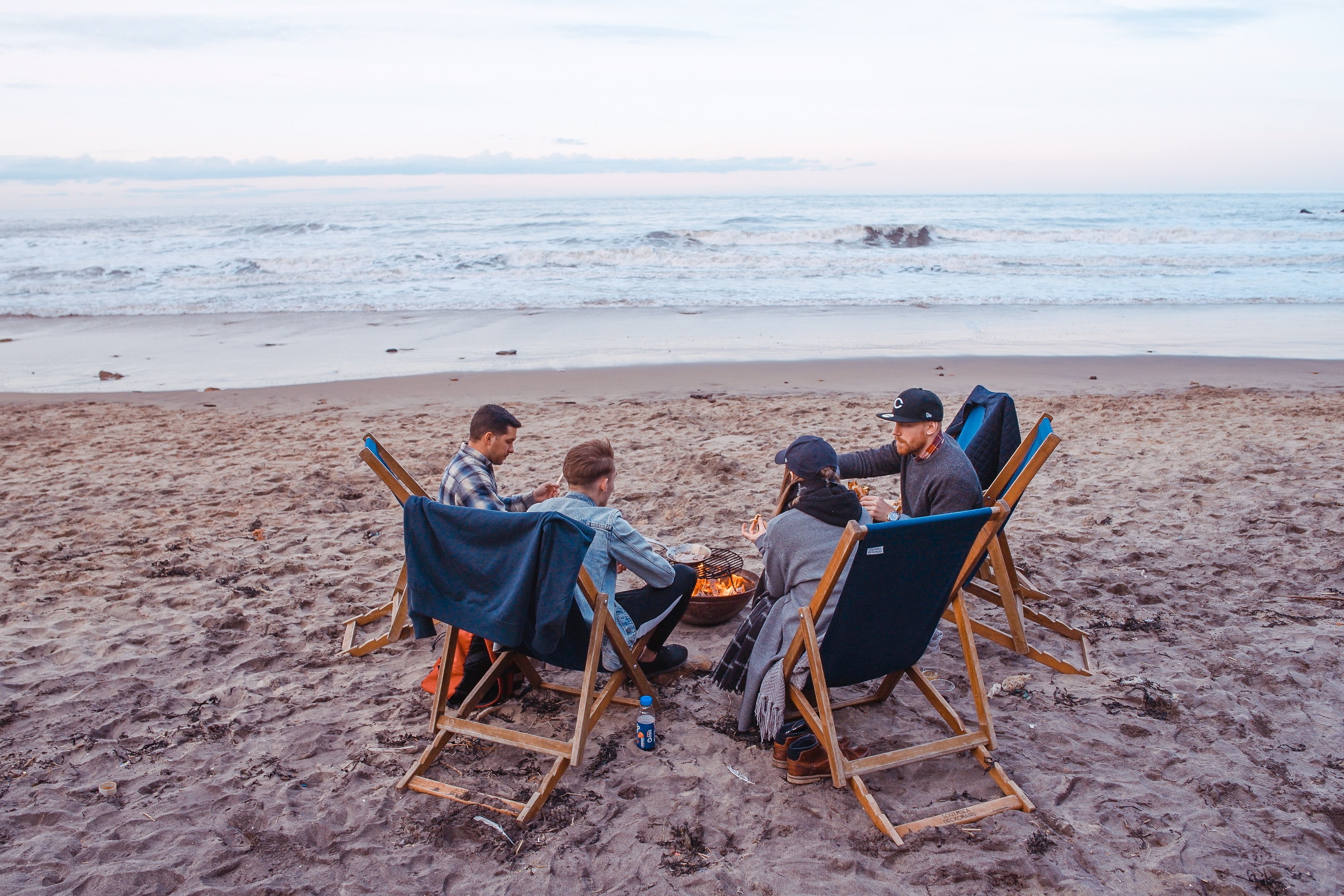 Three male and one female friend wit around a camp fire on a beach at dusk. They have prepared food and are eating together. While it's not always easy to invite non-believer friends to read the Bible with you, God will guide your time.