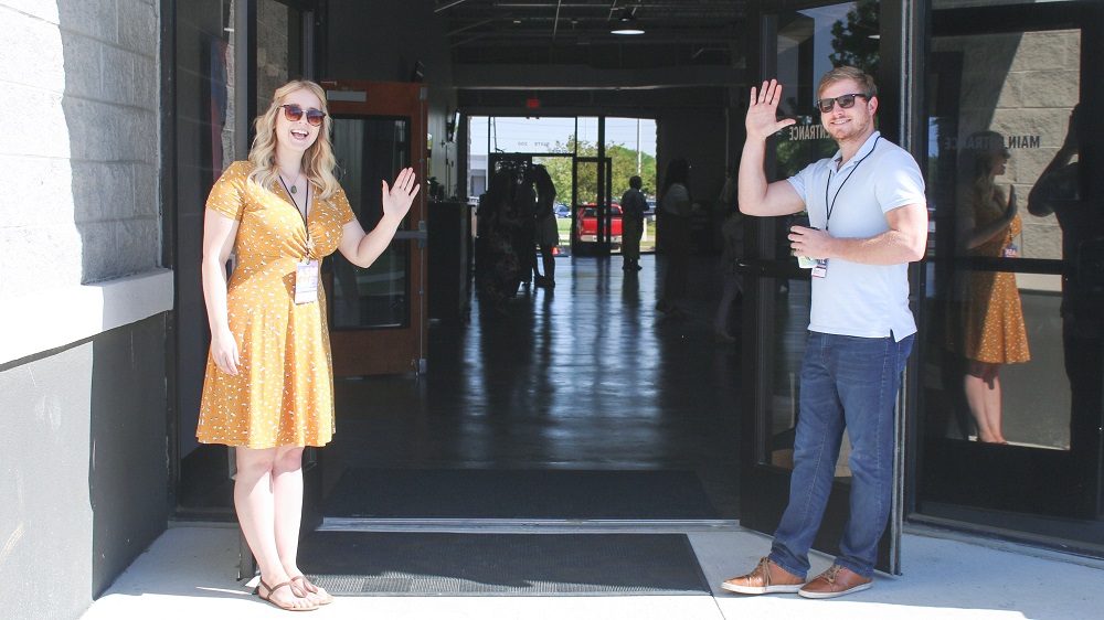 A man and a woman stand in the entrance to their church waving, ready to joyfully greet people as they enter. they are using their spiritual gifts to help in their church.