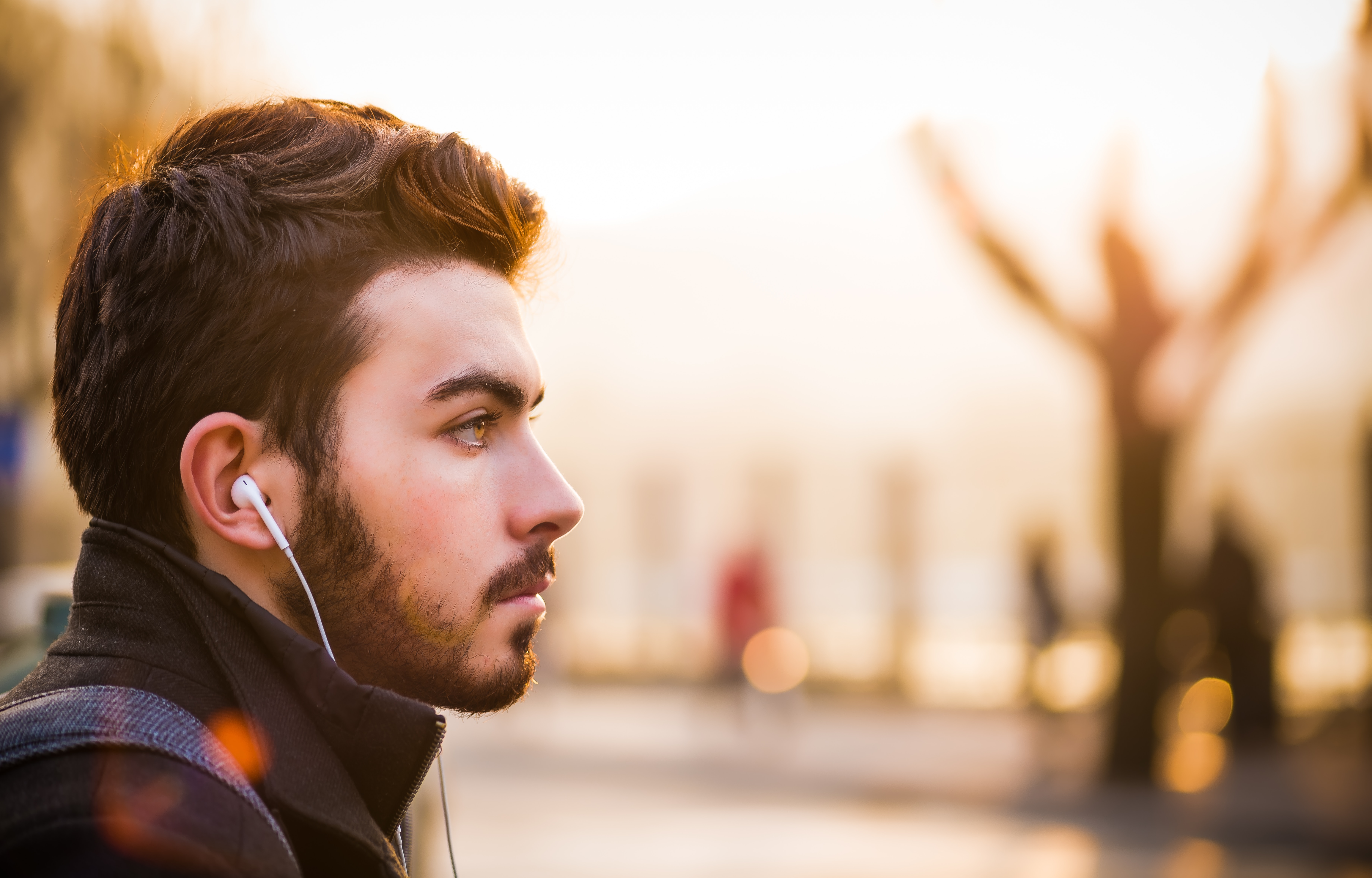 A young man is pictured in profile, with headphones in his ears and staring off into the distance. He is outside. He may be praying silently for his non-believer friends.