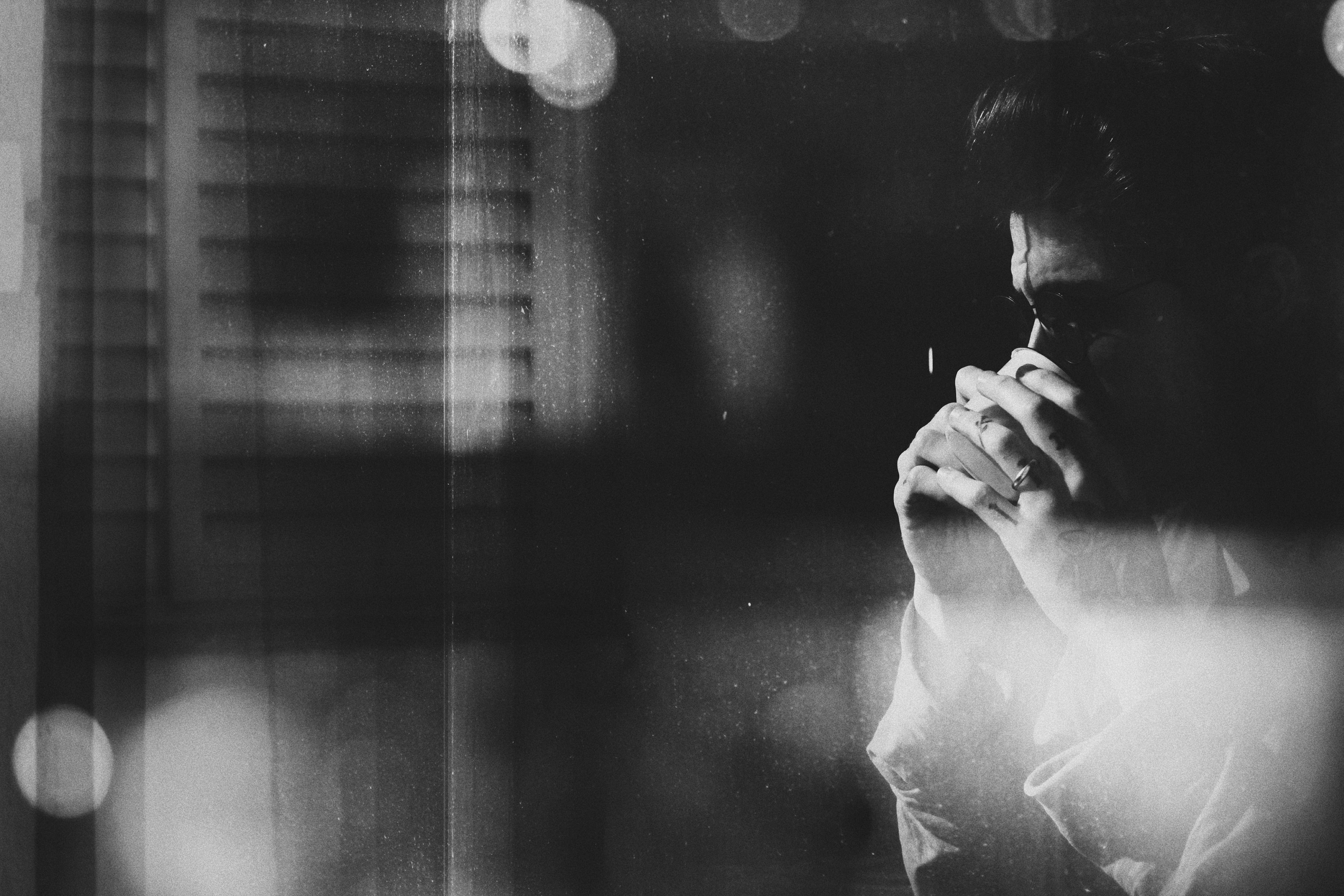 A man, in a black and white shot, sites beside a window with a bind over it, with his hands held together in front of his face, as if he is praying for a non-believer friend.