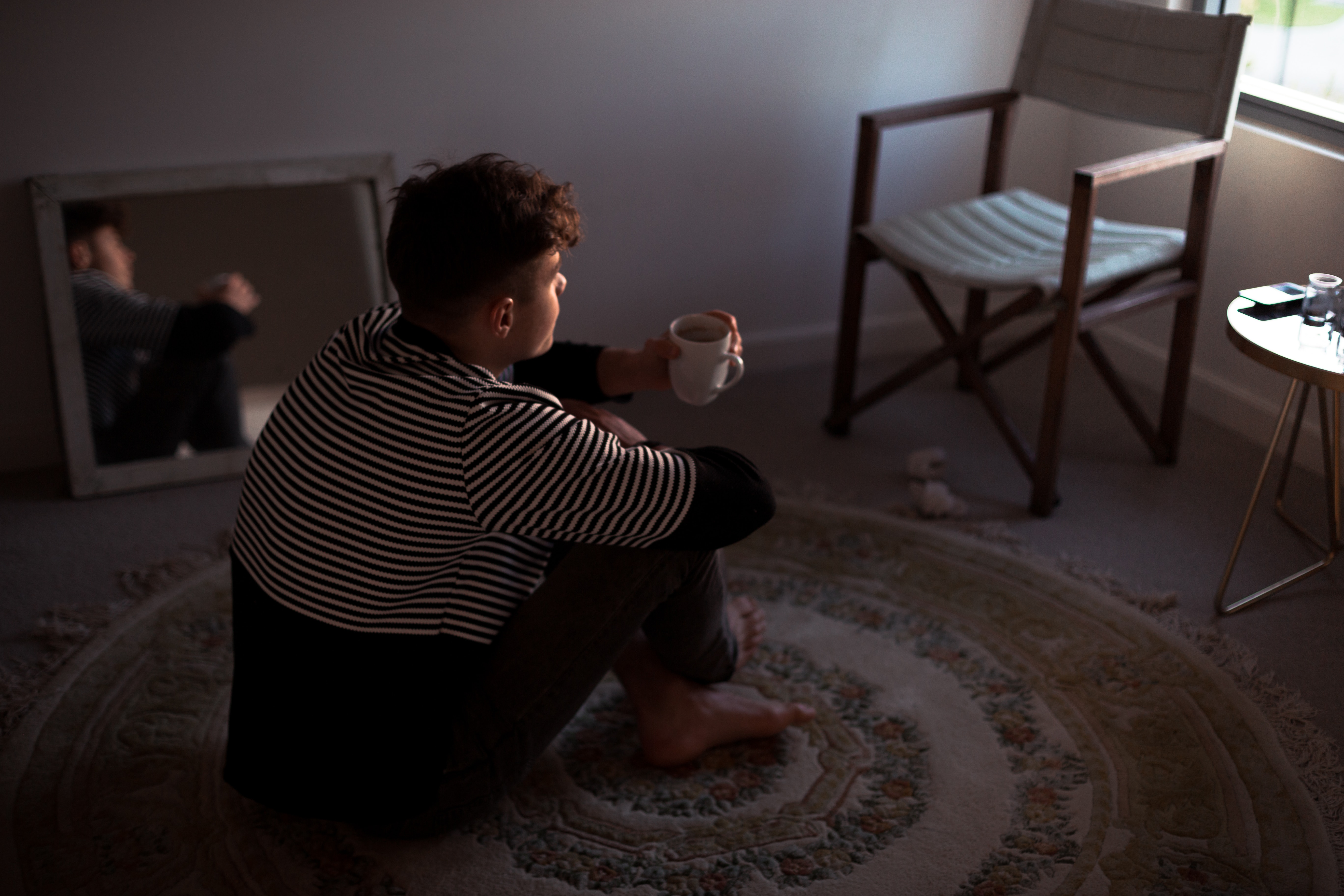 A young man sits barefoot on a circular rug with a mirror beside him and a chair and side table in front of him. He looks out of the window ahead while holding a cup and looking somewhat pensive.