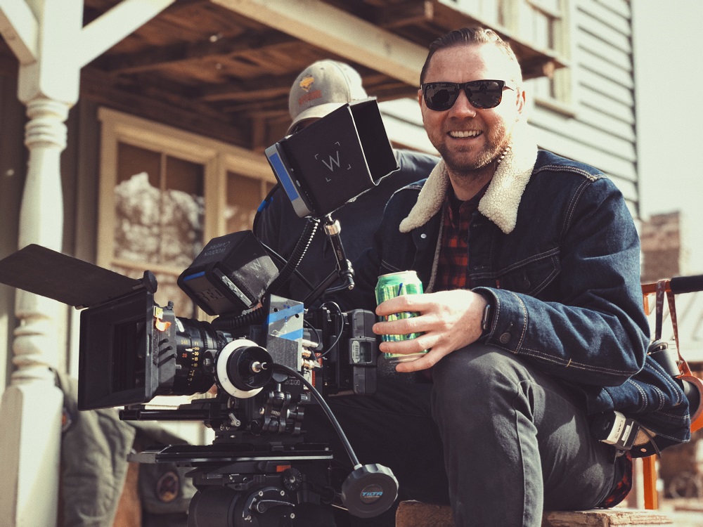 A man sits behind a professional film camera. He is holding a can of drink, wearing sunglasses and smiling. He uses his spiritual gifts to help create video content for his church.