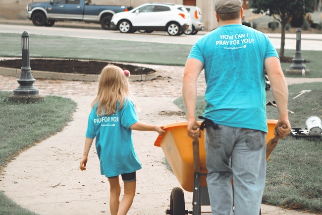 A father and daughter, wearing blue volunteer t-shirts, move a wheelbarrow as they use their spiritual gifts of helps to volunteer on a project.