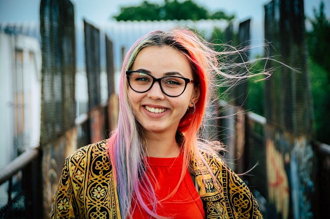 A young woman with multi-colored hair and trendy glasses stands smiling at the camera. She clearly finds joy in using her spiritual gifts in the place where God wants her!