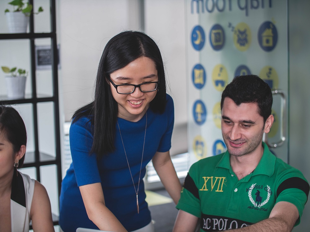 A woman stands over a man, helping him with some work. They're both smiling and look like they're in an office or class setting. She is using her spiritual gifts to teach and help.