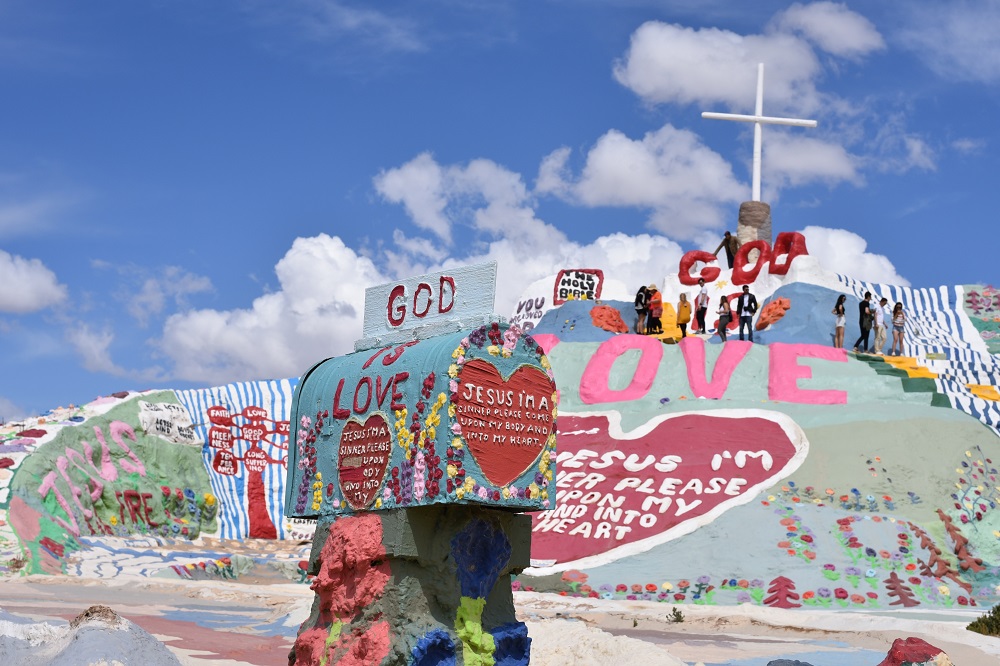 We see a large hill covered in a colorful mural with a tall white cross on it and a painted mailbox in the foreground, all proclaiming the love of God and Jesus. The artist who created this extraordinary piece is putting their spiritual gifts on full display!