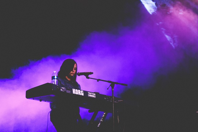 A woman plays on an electric keyboard on stage with smoke behind her lit in purple. She is using her talent and skills to inform her spiritual gifting and find the very best place for her to exercise them.