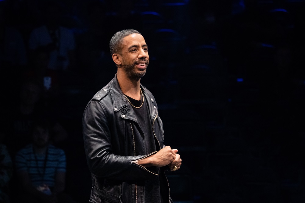 A manin a leather jacket, with a headpiece microphone, speaks from a dark stage. He is using his spiritual gifts of teaching and exhortation.