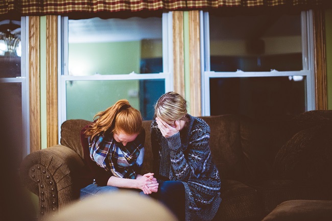 Two young woman sit on a couch with their heads bowed in prayer. Faith, healing and mercy are all spiritual gifts they may be using in this moment.