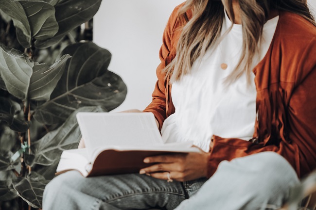 A woman sits with a Bible open on her lap and she leaves through the pages. A large green plant is next to her. She is devoting time to God's Word in order to abide in Christ.