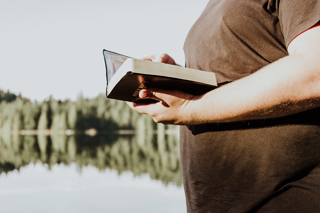 A Bible reader in a brown t-shirt stands next to a still lake with their Bible open and the sun illuminating them. To abide in Christ is to walk close by Him, and by reading the Bible daily we can remain close to Him.