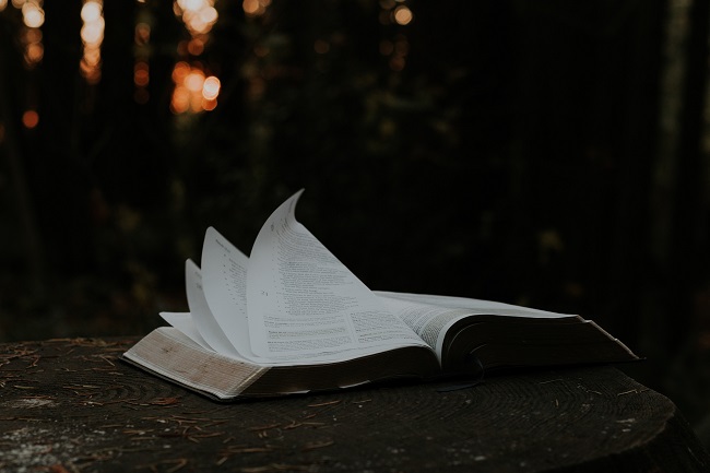 An open Bible lays on a wooden table in what appears to be a back yard. it's pages flutter in the wind. Bible study helps us to keep learning about Jesus and becoming more like Him.