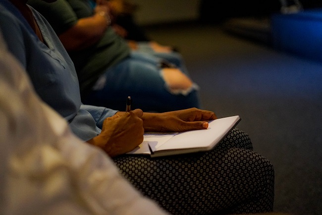 We see a man's hands writing in a book as he sits listening to a lecture with people on either side of him. He is learning in order to follow Jesus closely.