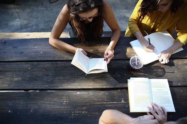 Three friends sit around a wooden bench with books, making notes in them, studying the Bible to keep learning about Jesus.